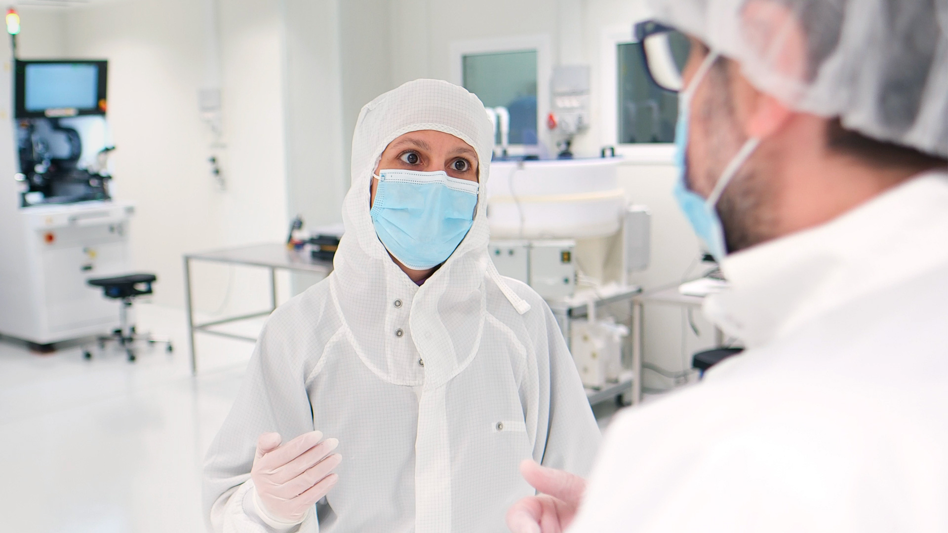 Technicians wearing clean room garments and protective equipment discussing activities inside an ISO 6 clean room.